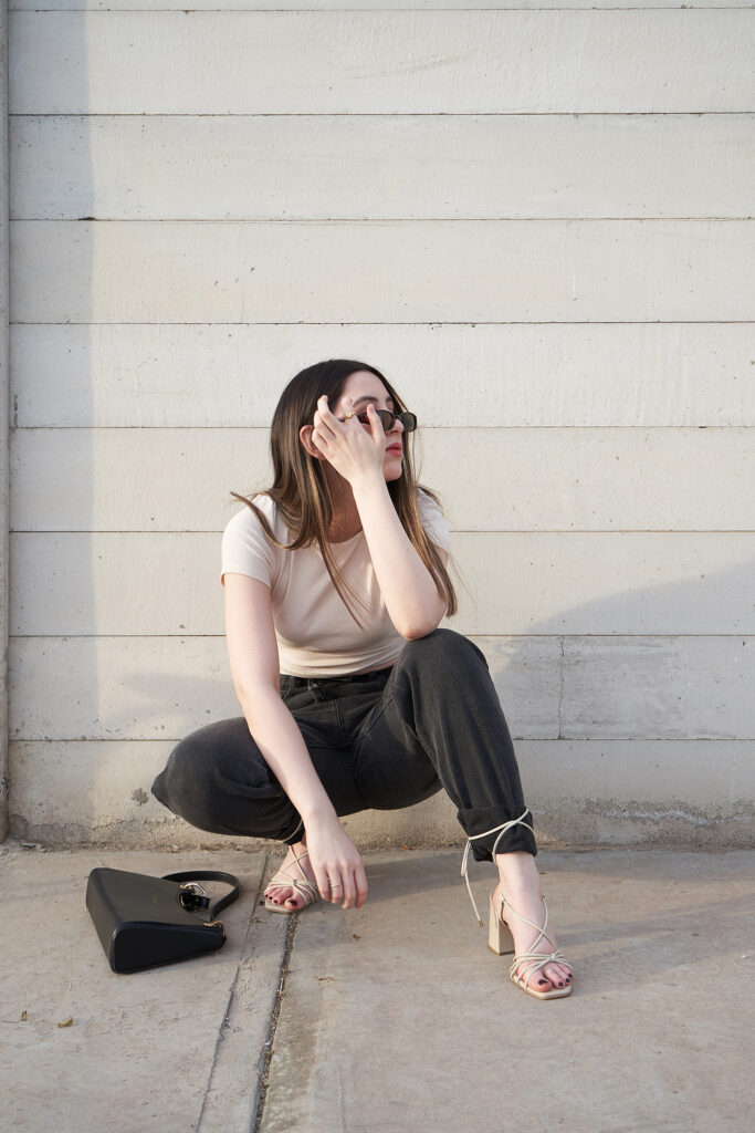 Gabriela Inzunza for Avice crouching against a white wall wearing a beige top black trousers and strappy sandals with a black handbag beside her.